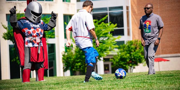 Two people kick a soccer ball together on the quad while Barry watches.