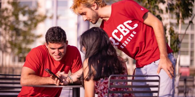 Three RCBC students sitting outside at a table looking at a phone