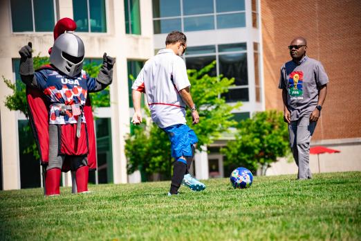 Two people kick a soccer ball together on the quad while Barry watches.