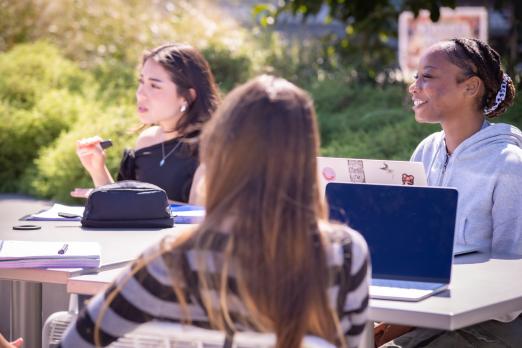 Three students sitting outside at a patio table with laptops and notebooks, talking together in a sunny campus quad.