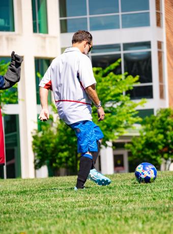 Two people kick a soccer ball together on the quad while Barry watches.
